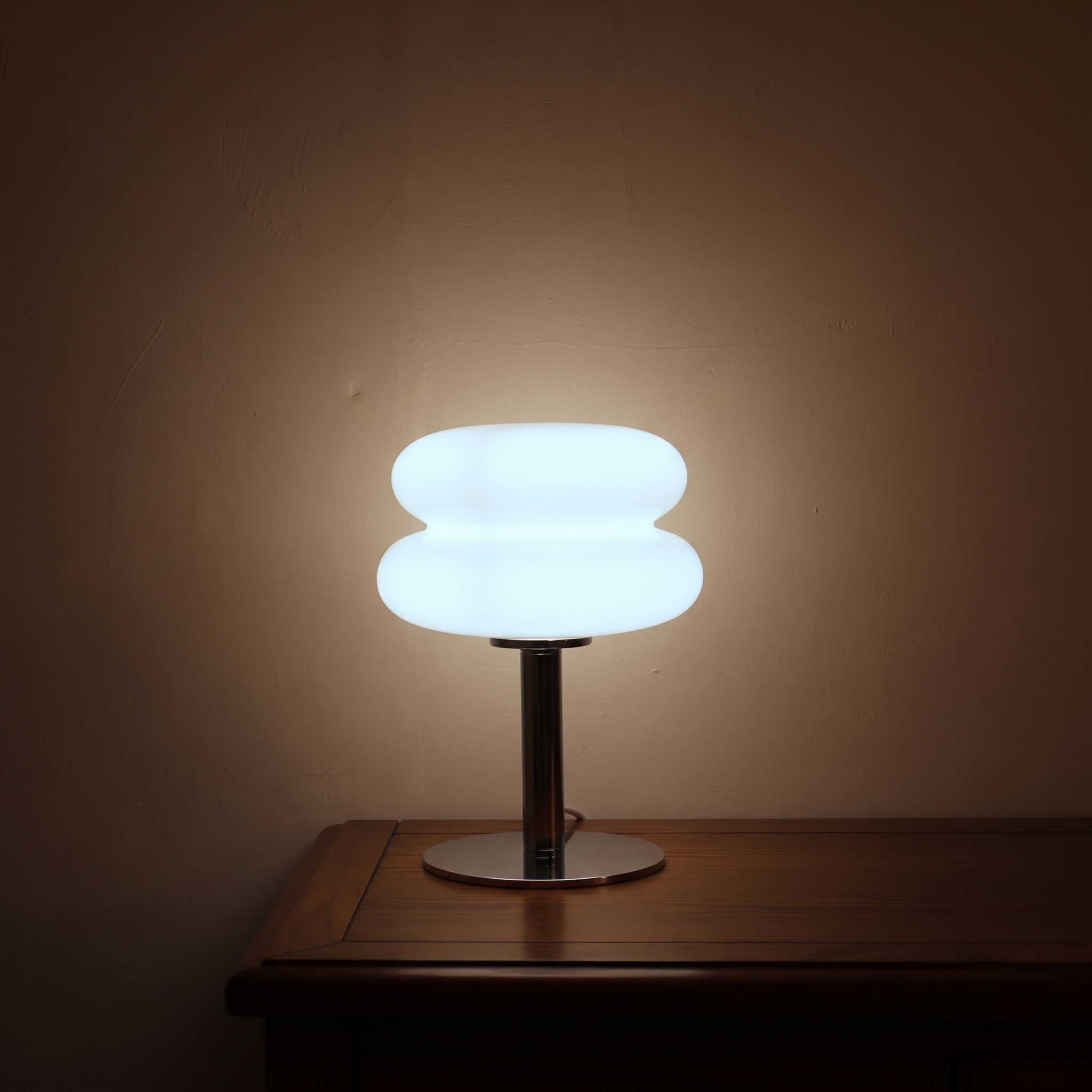 White Macaron Glass Table Lamp lit in a dark room on its cool white setting, casting pale light up a plaster wall.