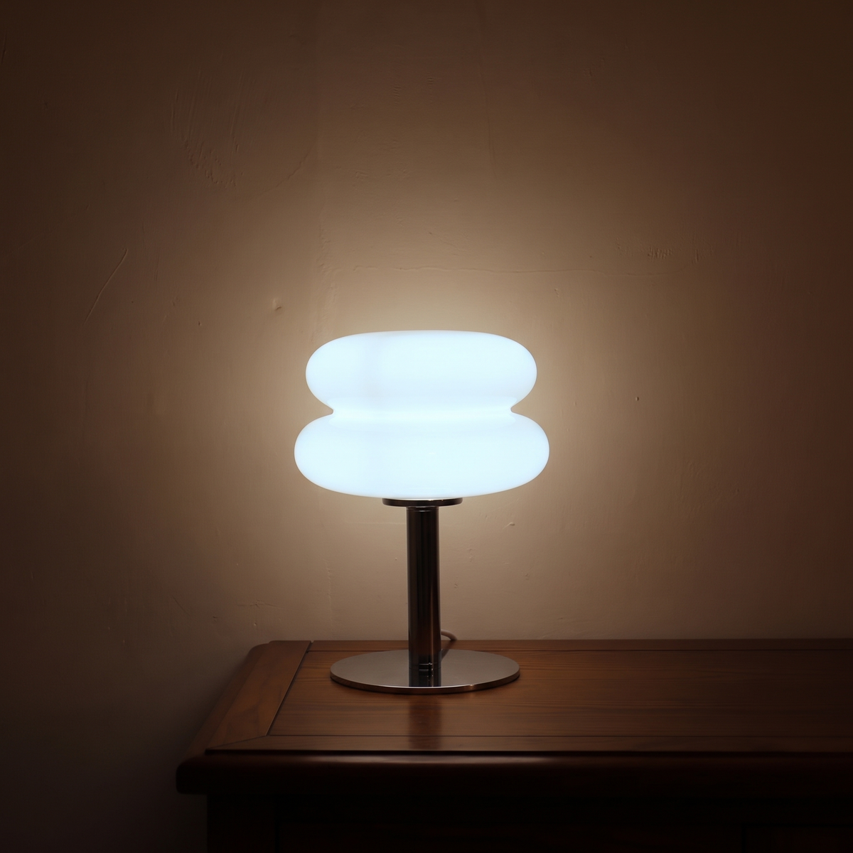 White Macaron Glass Table Lamp lit in a dark room on its cool white setting, casting pale light up a plaster wall.