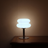 White Macaron Glass Table Lamp lit in a dark room on its cool white setting, casting pale light up a plaster wall.