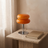 Orange Macaron Glass Table Lamp switched off on a travertine nightstand beside a hardback book, showing its 11 inch height.