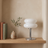 White Macaron Glass Table Lamp switched off on a white oak shelf in daylight, beside two books and a ceramic vase.