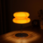 Orange Macaron Glass Table Lamp lit in a dark room, warm amber light glowing through the glass and pooling across a walnut surface.