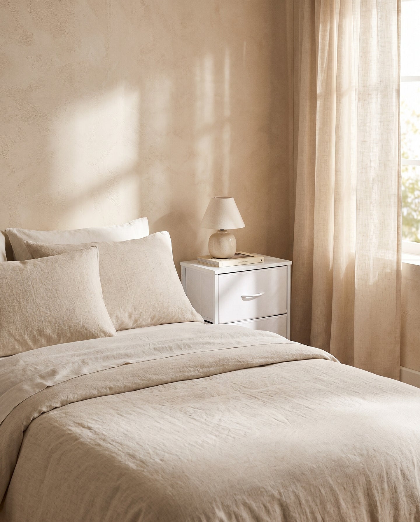 Fabric drawer nightstand in white, styled beside a bed with oatmeal linen bedding in a warm plaster-walled bedroom