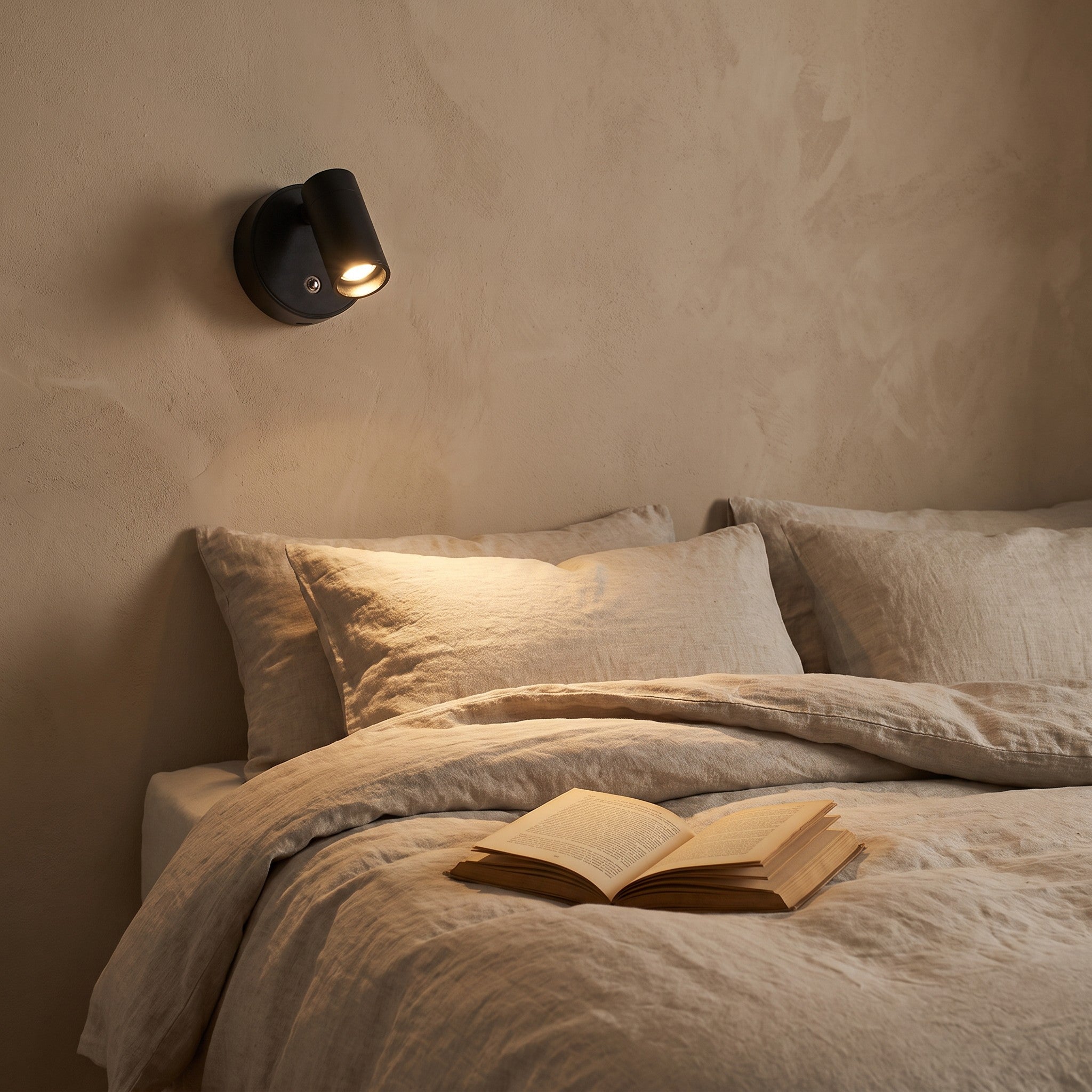 Reading light lit beside a bed with linen bedding and an open book