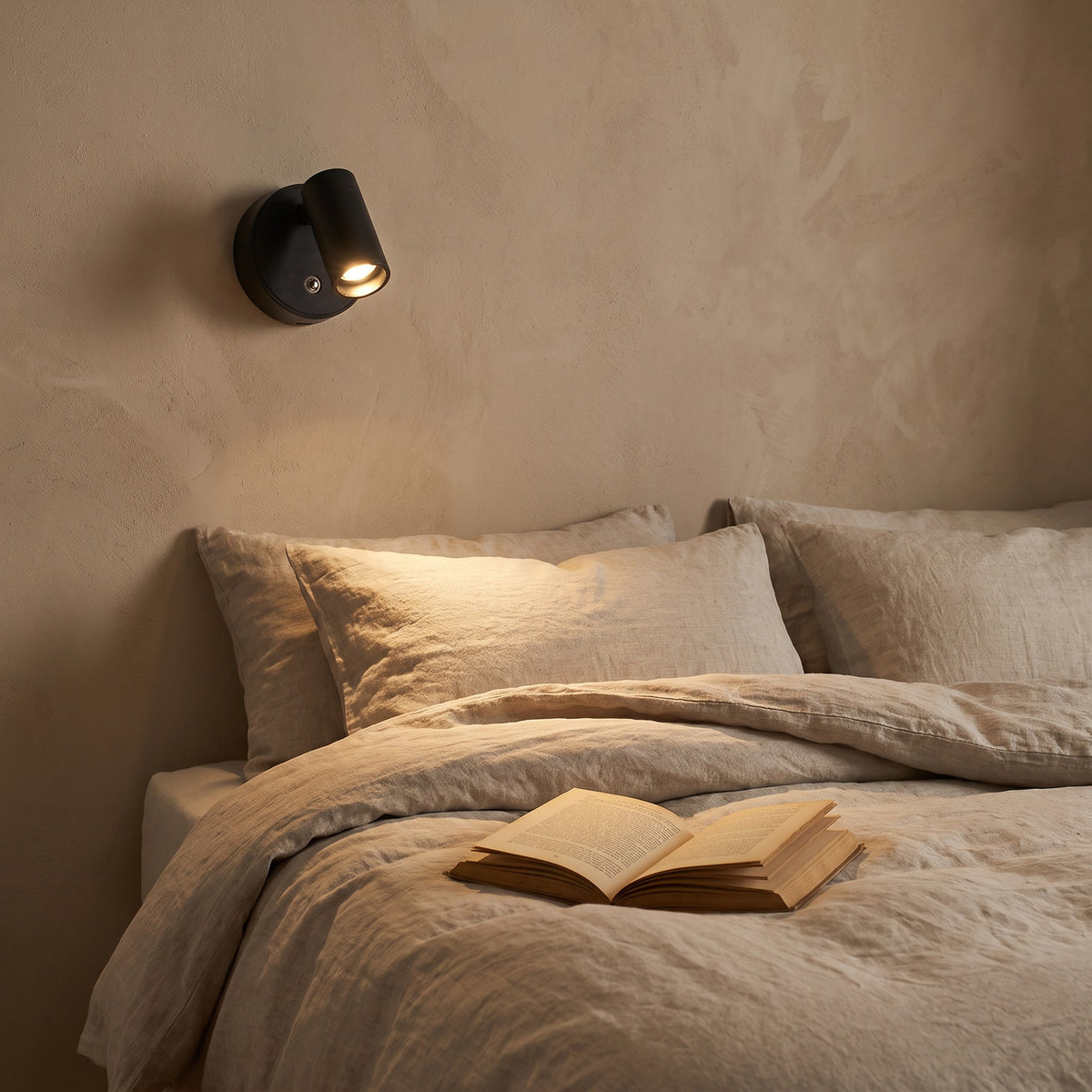 Reading light lit beside a bed with linen bedding and an open book